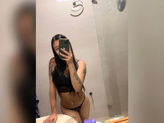 LuciaCalixto, con cabello negro largo, vestida con un sujetador y tanga de encaje negro, sosteniendo un teléfono en un selfie frente al espejo, mostrando tatuajes en el brazo y el costado, y un piercing en el ombligo..