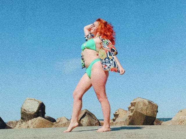 SpicyGrace, com cabelo ruivo, biquíni verde e camisa aberta, em pé sobre rochas, descalça, com a mão no cabelo, mostrando o bumbum e os seios, sob um céu azul claro..