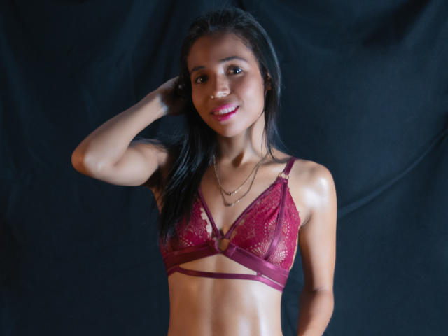 LaylaBrown, avec une peau foncée et des cheveux noirs, porte un soutien-gorge en dentelle rouge, a de petites seins et une silhouette mince, elle sourit avec du rouge à lèvres rose, une main dans les cheveux, fond noir..