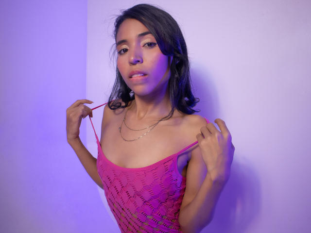 Layla Browns, con la pelle marrone media, i lunghi capelli neri e il seno piccolo, indossa un abito rosa trasparente all'uncinetto, con spalline, davanti a un muro illuminato di viola, e guarda verso la fotocamera..