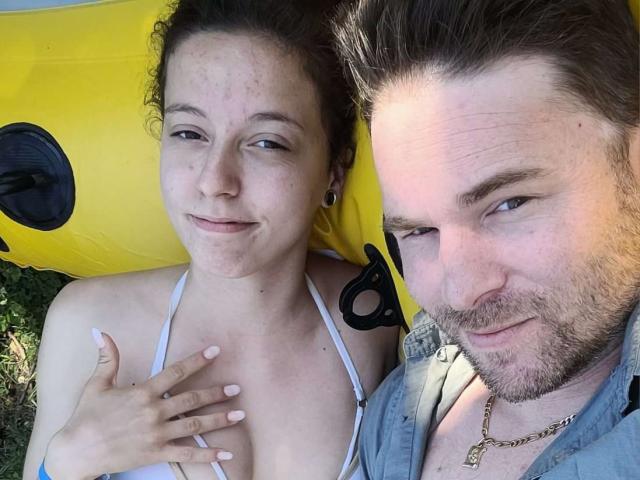 LilaXTom with freckles, small tits, white top, hand on chest, smiling, and Tom with beard, short brown hair, gold chain, gray shirt, yellow inflatable, slightly smiling.