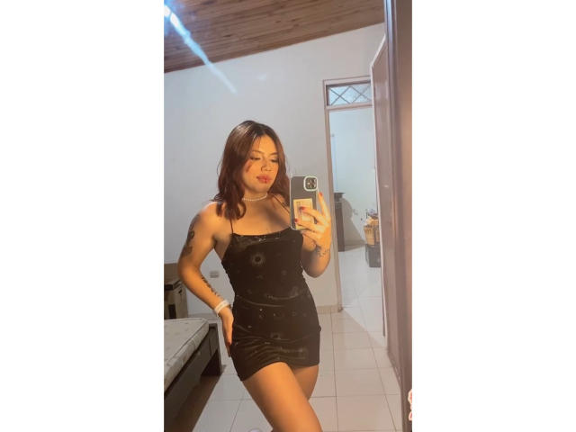 SophiaMarx in a tight black dress, standing in a mirror selfie, showing her ass and small tits, with brown hair, a bracelet, and a confident smile, holding a phone.