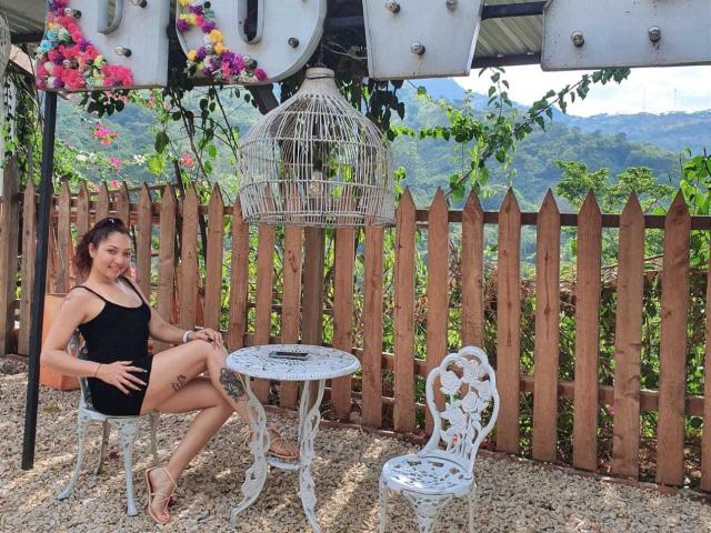 KatrinaBello in a black dress, sitting with a white, ornate chair and table, her ass visible, next to a birdcage, wooden fence, and a mountain view in the background.
