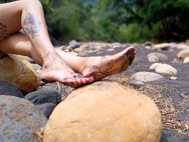 KatrinaBello's dirty feet with red toenails, crossed ankles, showing a colorful tattoo on her left leg, sitting on a rocky, sandy beach. Her right foot has mud on it.