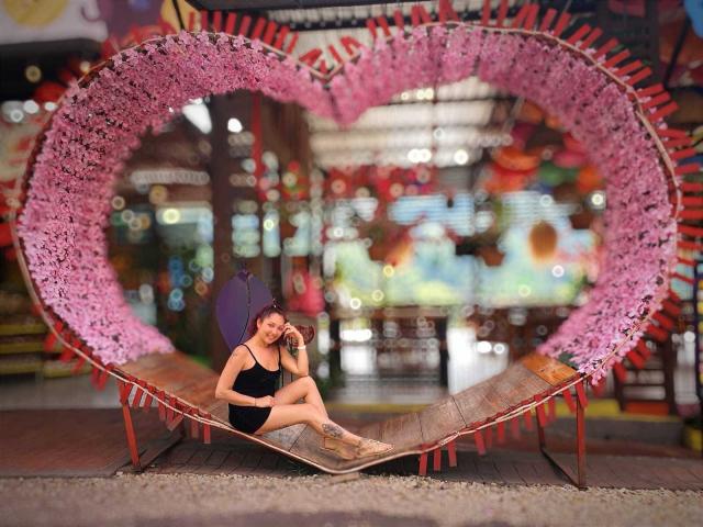 KatrinaBello assise sur un banc rose en forme de cœur, avec des bords ornés de fleurs, portant un débardeur noir et un short, regardant son téléphone, avec un parapluie violet derrière elle..