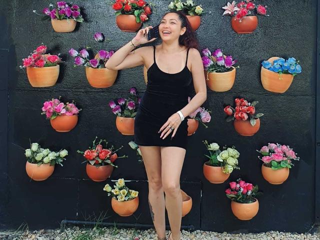 KatrinaBello stands smiling, wearing a tight black dress, showing her tits and ass, with curly red hair, against a black wall with orange flower pots.