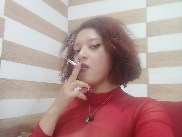 VioletaSwitch with curly red-brown hair, wearing a red sheer top, has a finger near her purple lips, a heart necklace, and a serious look, against a striped wooden wall.