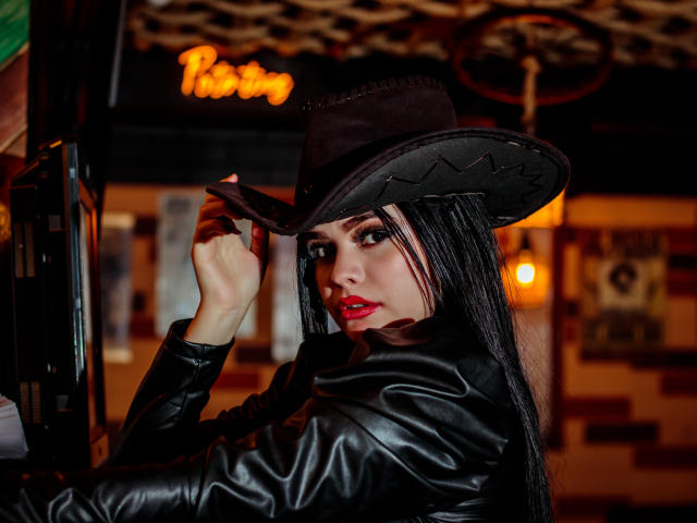 BrithneyLee, avec ses longs cheveux noirs, son rouge à lèvres rouge et sa veste en cuir noir, portant un chapeau de cowboy marron, le touchant avec sa main droite, dans un bar faiblement éclairé..