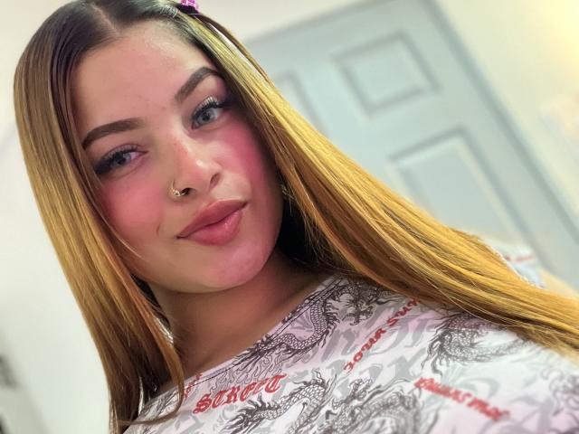 ShantalSummer with long blonde hair, pink blush, and nose ring, wearing a white patterned shirt, puckering her lips in a selfie. Her tits are slightly visible under the shirt.