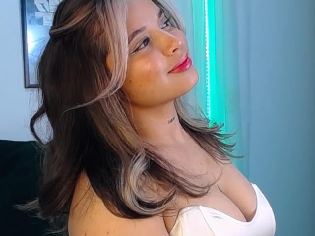 KateZoe with shoulder-length brown hair and blonde highlights, wearing a white strapless top, showing her tits and smiling with red lipstick, blue light in background.
