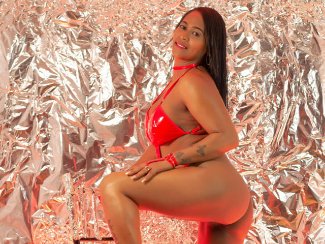 BeckyVega with dark hair, red latex bikini, showing her ass, sitting on a stool, smiling, tattoo on her right wrist, silver foil background, red light.