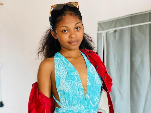 LucresiaAdel with dark skin, brown eyes, and curly black hair wearing a blue paisley halter top, red jacket off shoulders, and sunglasses on head, smiling softly.