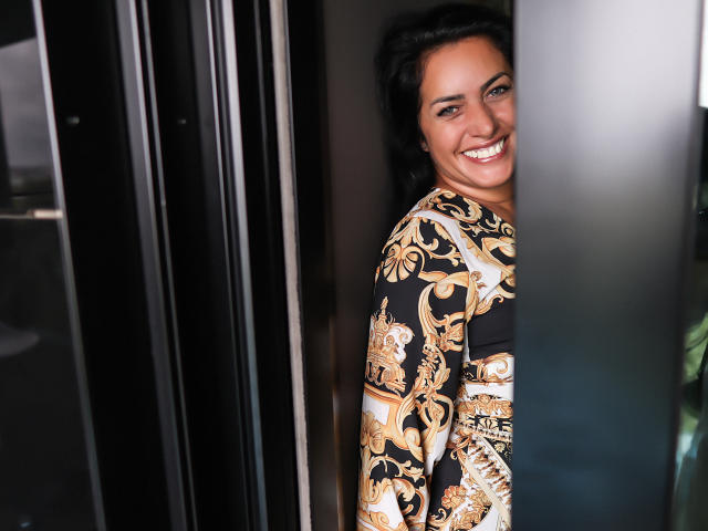 AnelisaReys, avec ses cheveux sombres et son sourire, jette un coup d'œil derrière une porte, vêtue d'une robe noire et dorée à motifs floraux, qui dévoile légèrement sa poitrine..