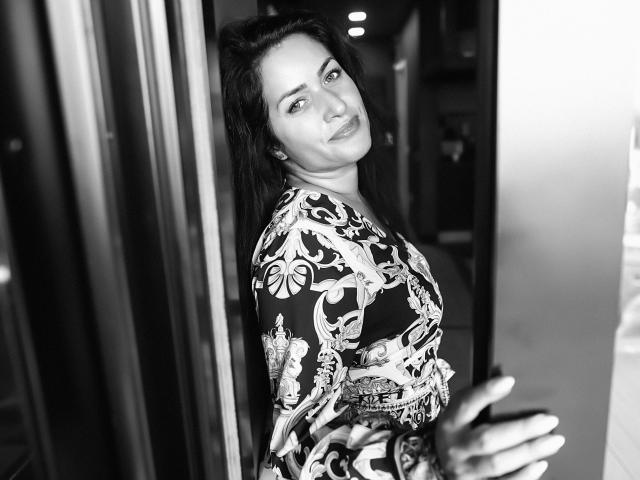Fotografía en blanco y negro de AnelisaReys con cabello oscuro, vestido con estampado floral, de pie en un marco de puerta, girando ligeramente la cabeza, sonriendo, con una mano en la puerta, mostrando sutilmente sus pechos con el vestido..