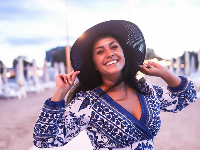 AnelisaReys with dark hair, smiling, wearing a blue and white floral top, and a big black hat, holding it with both hands, on a beach at sunset.