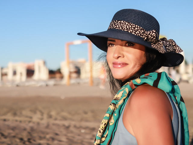 AnelisaReys, com pele morena, cabelos escuros, usando um chapéu azul com uma faixa marrom estampada e um cachecol verde, sorrindo para a câmera em uma praia ensolarada..