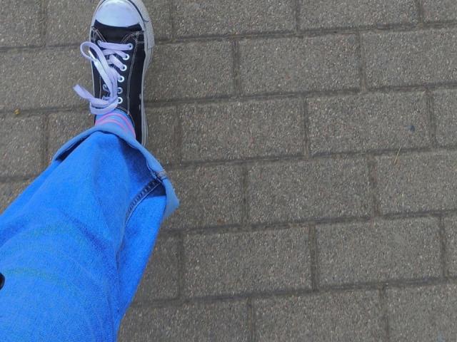 Zanoza's blue jeans and black sneakers with white laces, one leg slightly raised, showing a colorful sock pattern on a textured brick surface.