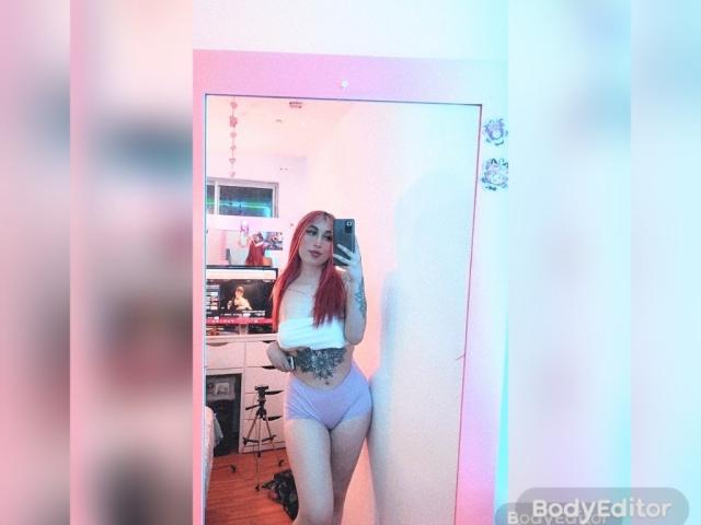 SammyViolet with red hair, white tube top, and purple shorts, showing off her tattooed stomach and large ass in a mirror selfie. She's holding a phone.