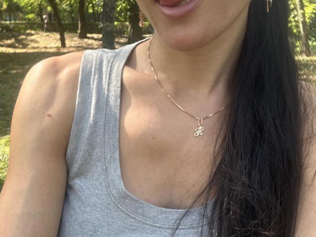BellaJulls con cabello negro largo, camiseta gris, busto pequeño, ligeras marcas de bronceado, una pequeña peca en el hombro, collar dorado, lengua afuera, luz del sol filtrándose entre los árboles..