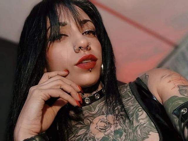 KaydeEbexley heeft zwart haar, tatoeages, rode lippenstift, een neusring, een lipring, een choker en rode nagels, met een hand in de buurt van haar gezicht, en ze kijkt er een beetje ondeugend uit..