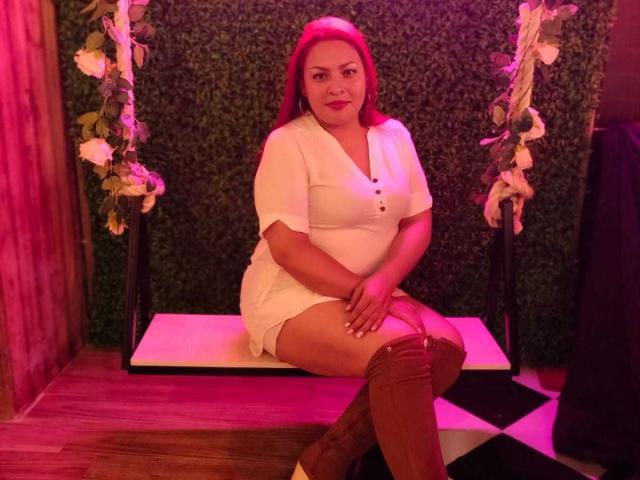 Adi69, avec des cheveux roux, une robe blanche et des bottes marron montantes, assise sur une balançoire, avec un éclairage rose et violet, un sol en bois et des décorations florales, regardant directement l'appareil photo..