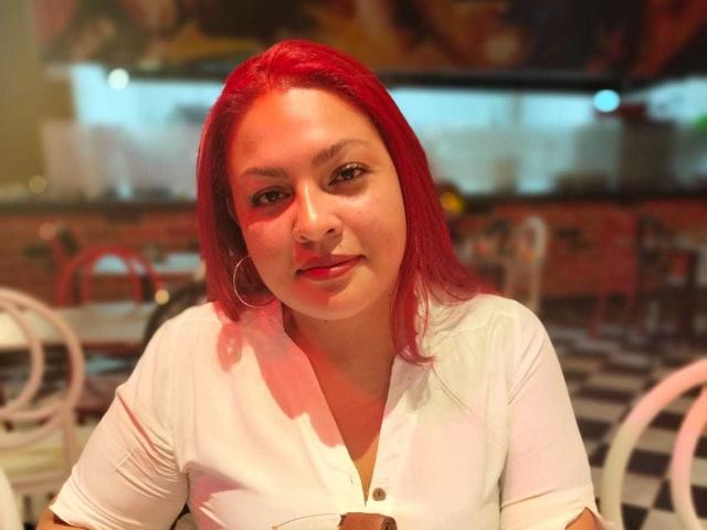 Adi69, com cabelo ruivo vibrante, camisa branca, pequenas argolas nos ouvidos e um leve sorriso, sentada em um restaurante, tem um rosto suave, levemente corpulento e uma atitude relaxada..