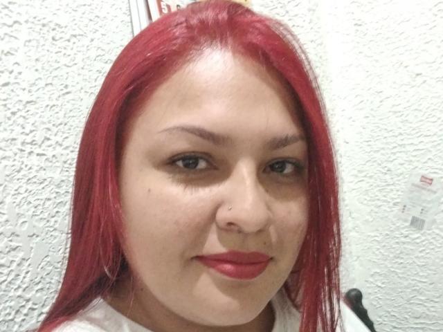 A Adi69 tem cabelo ruivo, pele clara, olhos verdes e um piercing no nariz. Ela está usando batom vermelho e uma camisa branca, com um leve sorriso. Seus seios não estão visíveis..