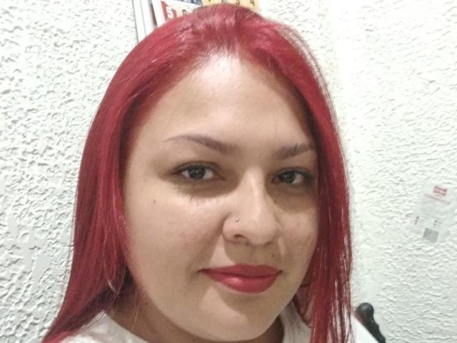 Adi69 hat rote Haare, helle Haut und ein kleines Nasenpiercing. Sie trägt roten Lippenstift und ein weißes Hemd. Ihr Gesichtsausdruck ist neutral mit einem leichten Lächeln..