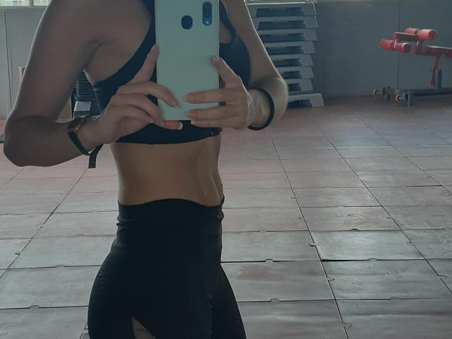 SeleneBrigth's toned abs and small tits are visible in a black sports bra, wearing black leggings, taking a mirror selfie in a gym with a blue phone.
