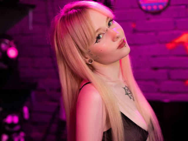 LaneyvilI, con lunghi capelli biondi, un anello al naso e tatuaggi, guarda verso l'alto con un leggero sorriso, indossando una maglia nera trasparente, con luci rosa e viola..
