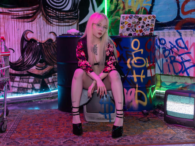 LaneyvilI mit blonden Haaren, Brille und pinken Strähnen, trägt einen Blumenbademantel, schwarze High Heels und hat Tattoos. Sie sitzt auf einer Kiste in einem mit Graffiti beschmierten Raum, die Beine auseinander..
