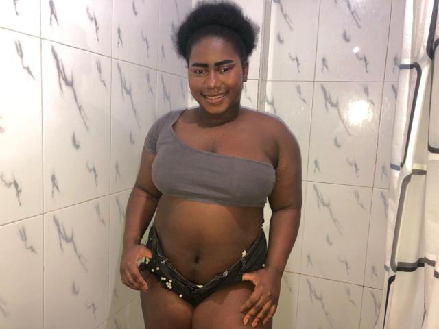 Dilay with dark skin, curly black hair, and pink eyeshadow, smiling, wearing a gray one-shoulder top and black sequined shorts, showing her tits and pussy.