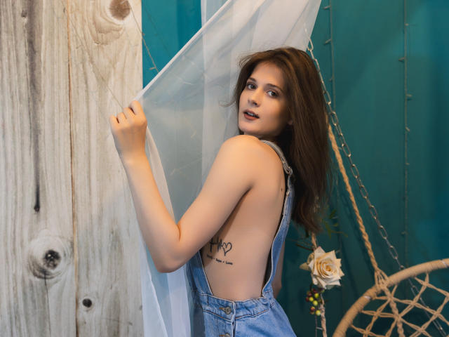 LeslieKatty with brown hair, wearing blue denim overalls, showing a side tattoo, holding a sheer white curtain, looking over her shoulder with a slight smile, showing her tits.