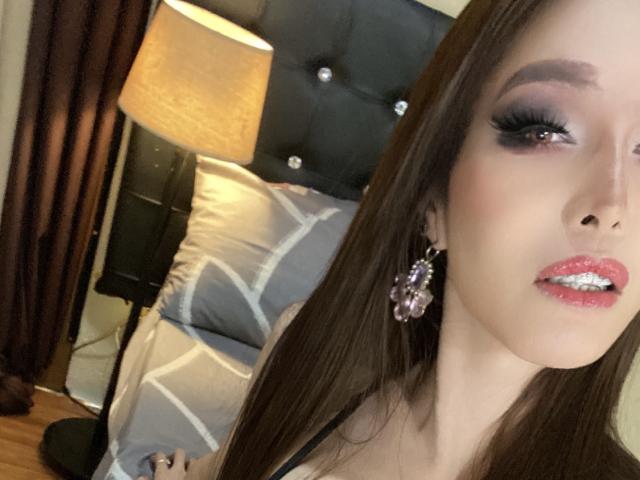 LaurentSuprema, con cabello largo y castaño, maquillaje de ojos morado, labial rosa brillante y aretes morados, mostrando su rostro con los labios fruncidos, acostada en la cama con un cabecero negro..