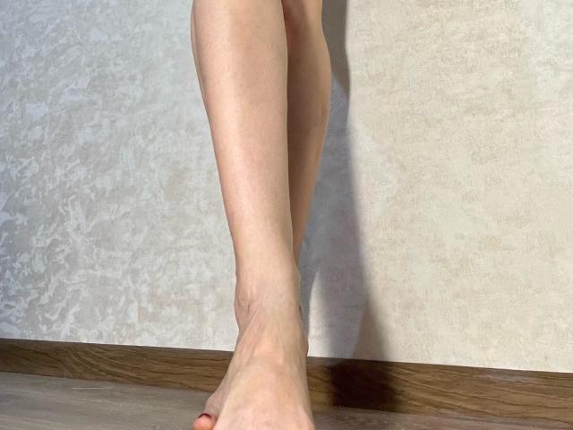 TheHottestBlonde's bare legs and feet with red-painted toenails standing on wooden floor, showing a slight shadow on textured white wall.