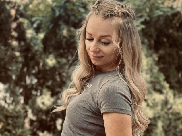 Elliette with long blonde wavy hair, braided crown, and gray shirt, looking down with a slight smile, her tits subtly visible, standing outdoors with green foliage in background.