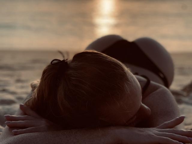Elliette, con il sedere in vista sotto delle mutandine nere, bacia la schiena lentigginosa di un uomo su una spiaggia sabbiosa al tramonto, con le mani appoggiate sulle sue spalle..