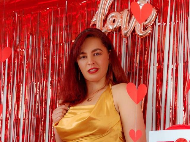 AmandaHotL with red hair, golden dress, red lipstick, holding dress, shiny silver background, red hearts, and silver "love" balloon. Boobs, tits, red hearts. Pretty, sexy, cute.