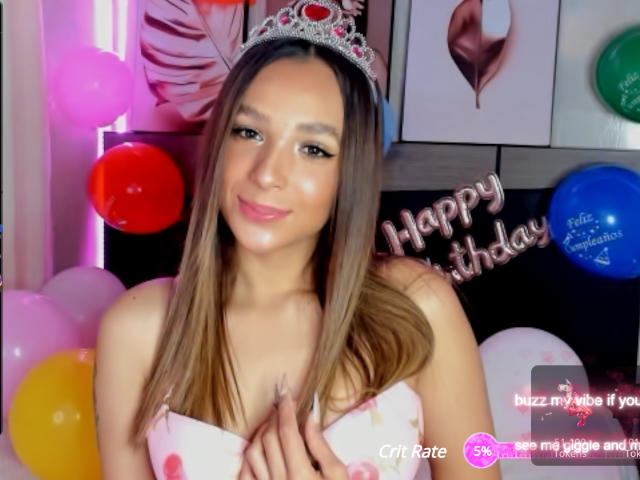 SarahWest with long brown hair, wearing a silver tiara, smiling, holding her small tits, wearing a white strapless top, surrounded by colorful balloons.