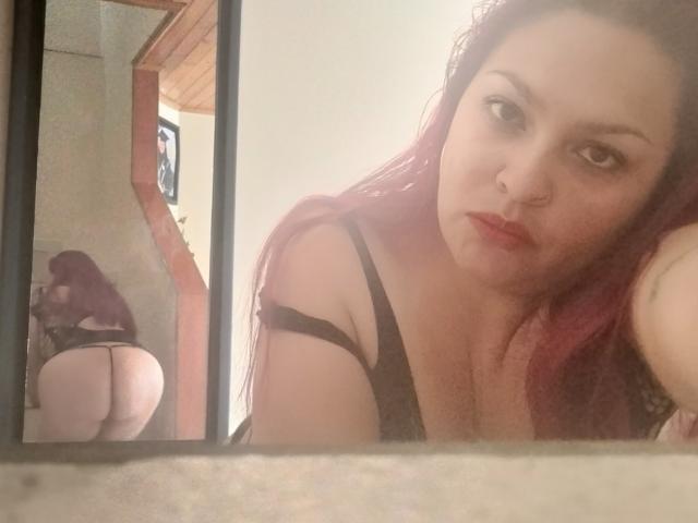 Adi69, con i capelli rossi, biancheria intima nera, un piercing al naso e rossetto rosso, mostra il suo sedere in un riflesso allo specchio. Il suo seno è piccolo e il suo sedere è prominente..