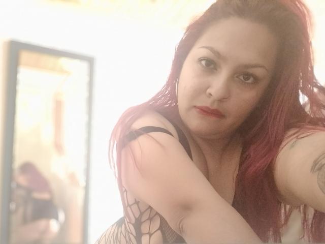 Adi69 with red hair, wearing black fishnet lingerie, has a tattoo on her arm, and is leaning forward with a serious expression, showing her tits in a soft-lit room.