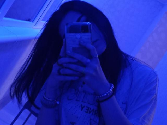 Zanoza, avec de longs cheveux foncés, un selfie pris devant un miroir légèrement bleuté, tenant un téléphone, portant une chemise, des bracelets et une bague, les yeux partiellement fermés, le téléphone couvrant partiellement son visage..