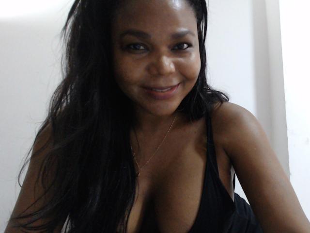 Una foto di BiggTitsx con la pelle scura, lunghi capelli neri e un sorriso, che indossa una maglia nera, scollata, che mette in mostra il suo seno grande e prosperoso. Indossa una collana d'oro..