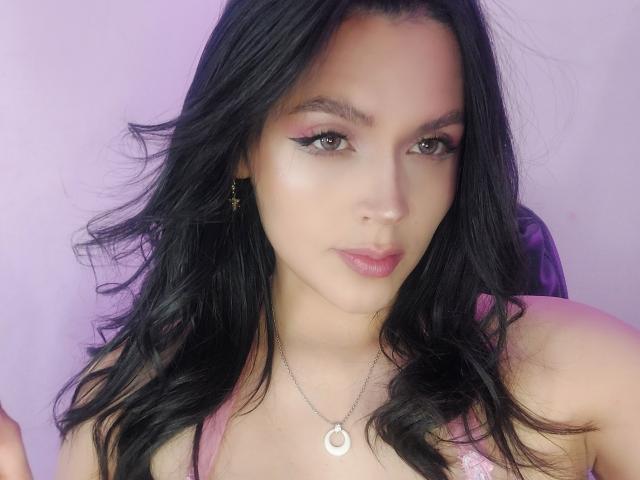 LinaStars with long black hair, green eyes, and pink lipstick, wearing a pink top, silver necklace, and star earrings, looking at the camera with a slight pout.