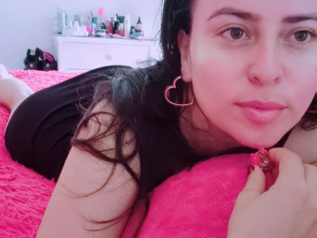 LindaSmith with pink heart earrings, lying on pink bed, black shirt, nose ring, holding ring, pink lipstick, dark hair, light skin, looking at camera, bedroom background.