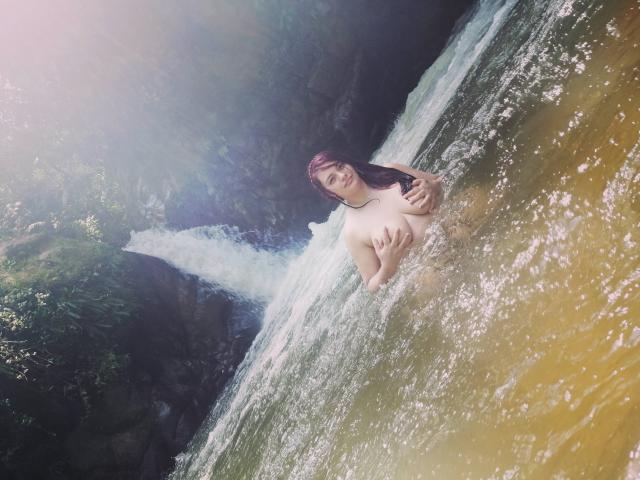 KatherinLatin, naked, with her tits covered by her hands, standing in a flowing waterfall, water splashing around her, looking at the camera with a slight smile.