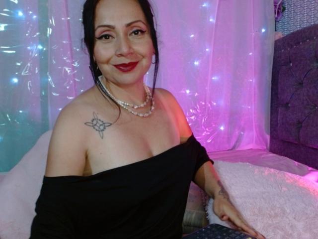 Zaria69 con una maglietta nera a spalle scoperte, rossetto rosso, collana di perle, tatuaggio sulla spalla destra, sorridente, che tiene una tastiera, luci viola e blu sullo sfondo. Il seno è visibile, la maglietta è tirata giù..