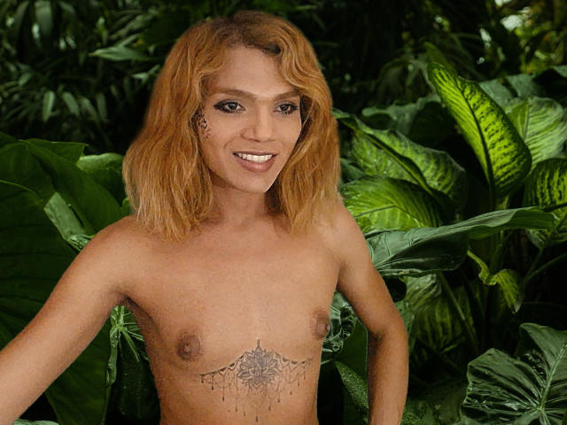 Topless TatyLorenX with orange hair, small tits, and a mandala tattoo below her tits, smiling in a lush green jungle. Her nipples are visible and she's attractive.