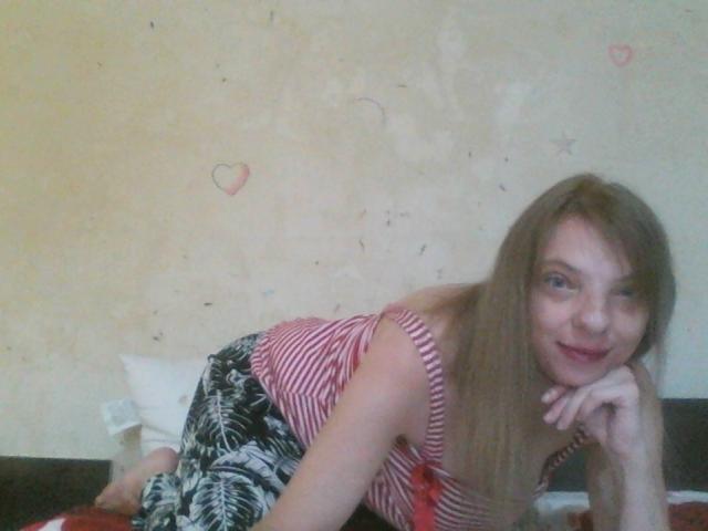 BlondyMarianne mit langen, braunen Haaren, einem gestreiften, rosa und weißen Oberteil, einem schwarz-weißen Rock mit Blattmuster, liegt im Bett, lächelt und hat ihr Kinn in der Hand, wobei kleine Brüste sichtbar sind..