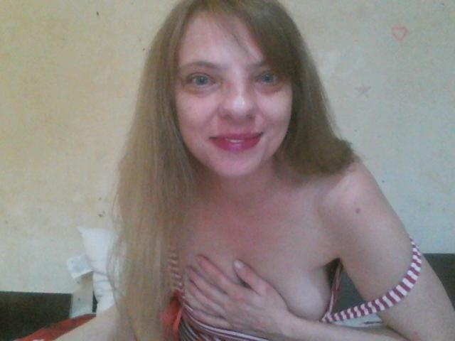 BlondyMarianne, com cabelos longos, batom rosa e olhos azuis, cobrindo os seios com a mão, vestindo uma blusa listrada em vermelho e branco, sorrindo para a câmera..